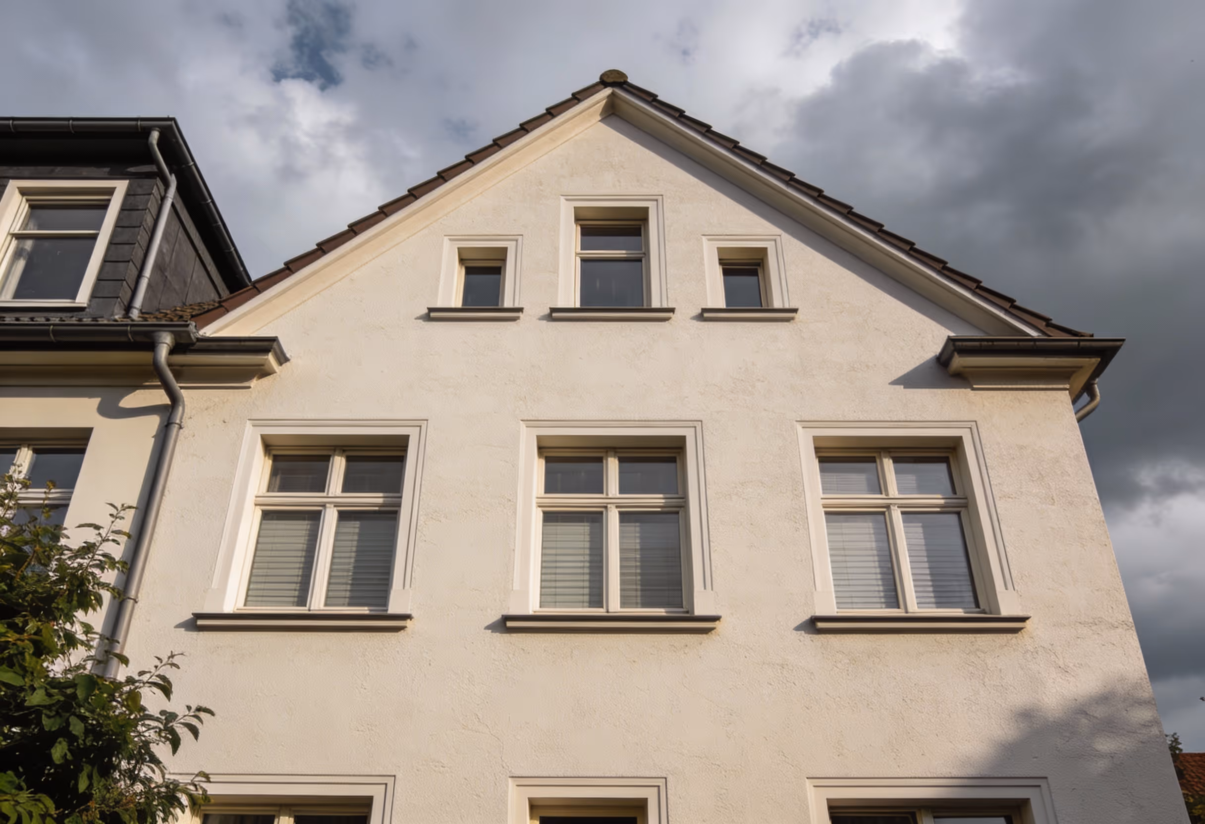 Ansicht der weißen Fassade eines Hauses mit mehreren Fenstern und bewölktem Himmel im Hintergrund.